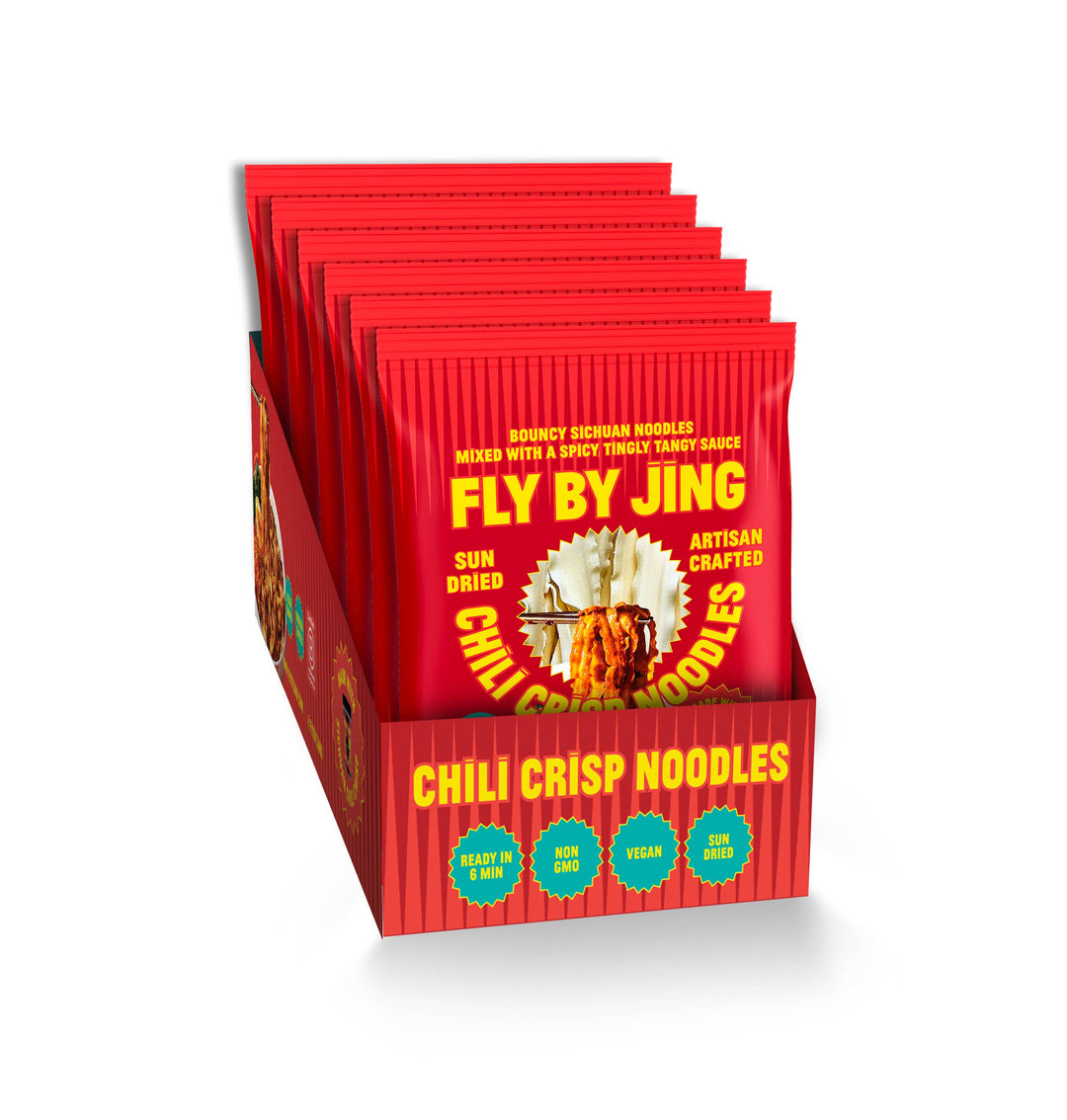 A retail display box filled with red individually wrapped packets of Fly By Jing Chili Crisp Noodles, shown on a clean white background.