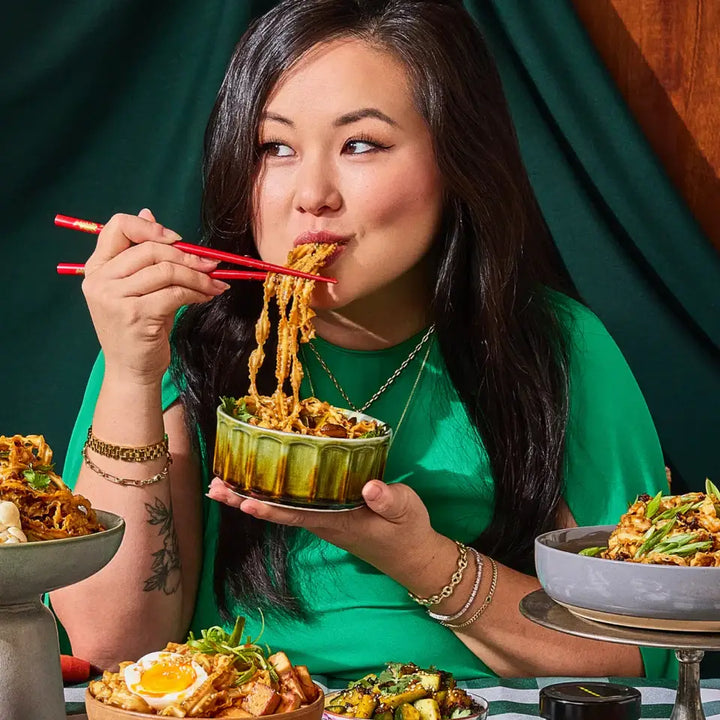 Woman eating noodles from a green bowl with chopsticks, surrounded by various dishes on a table.