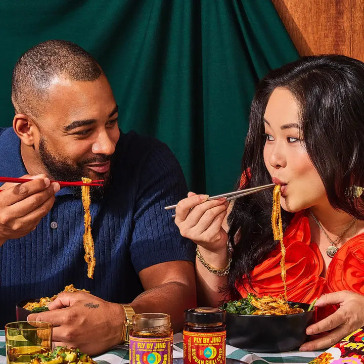 Two people enjoying a meal together with chopsticks, set against a green curtain background.