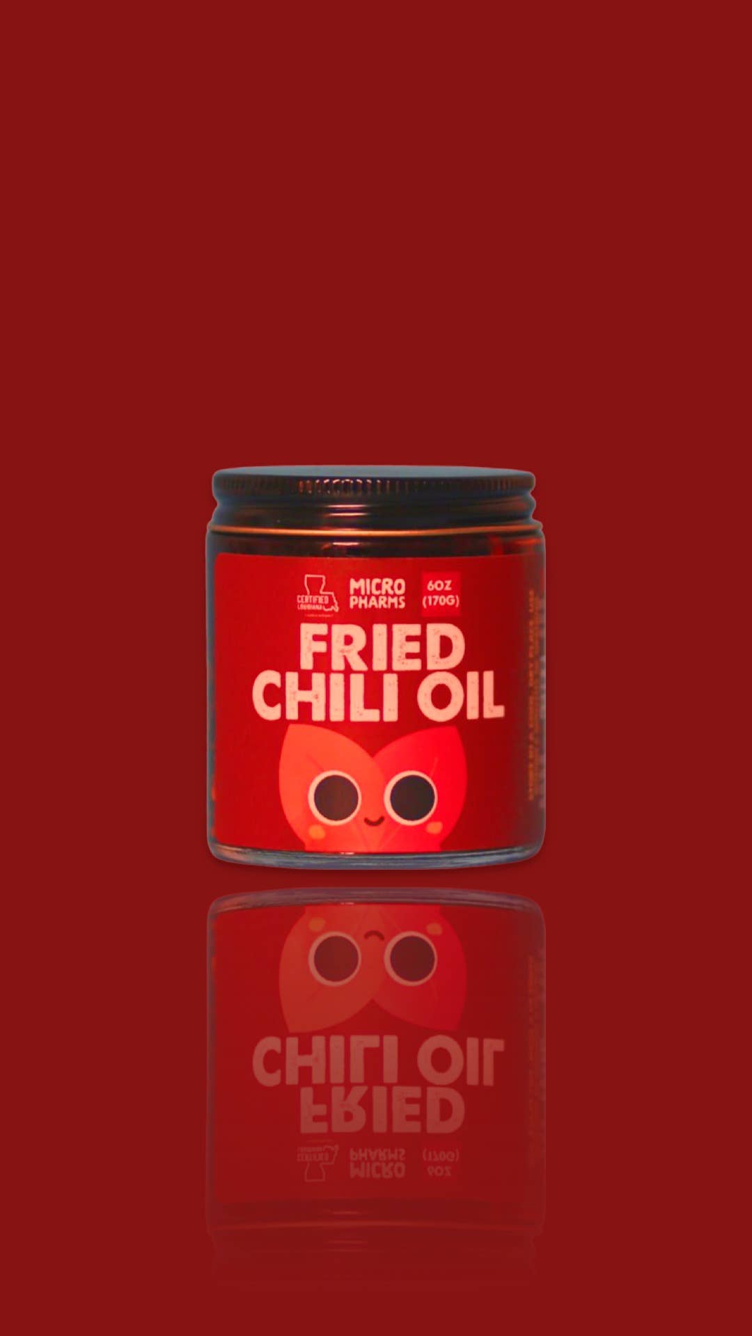 A jar of Micro Pharms Fried Chili Oil on a solid dark red background with a reflection below. The jar has a black lid and a red label featuring a cartoon chili face and white text.