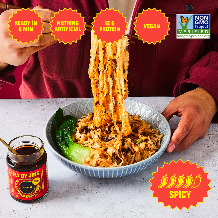 A bowl of Fly By Jing Vegan Chicken Noodle Soup is being lifted from the bowl by a person wearing a burgundy hoodie. The image features several promotional badges, including &
