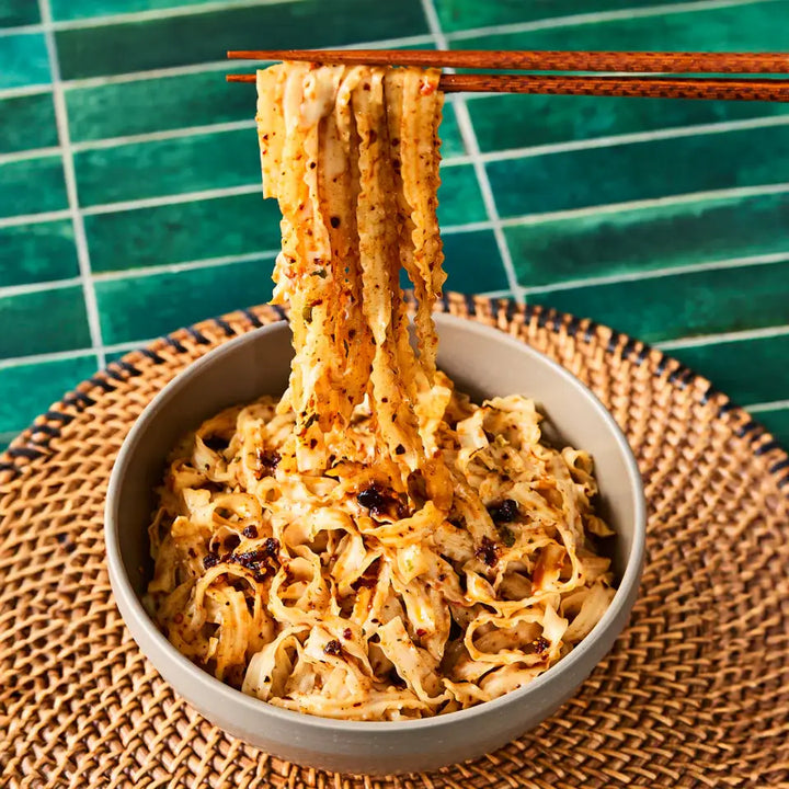Noodles being lifted with chopsticks from a bowl on a textured surface with a green tiled background