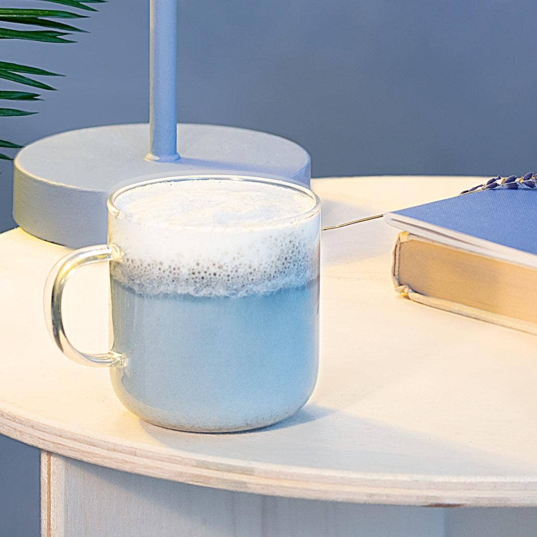 Glass mug with a frothy beverage on a table next to a book and lamp.