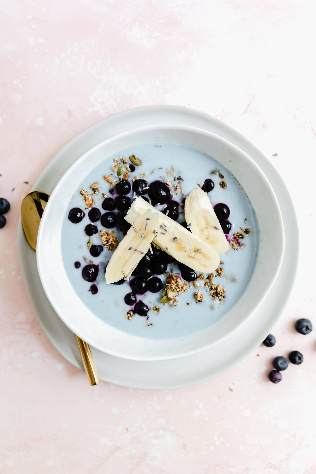 Blue smoothie bowl with bananas, blueberries, and granola on a pink background