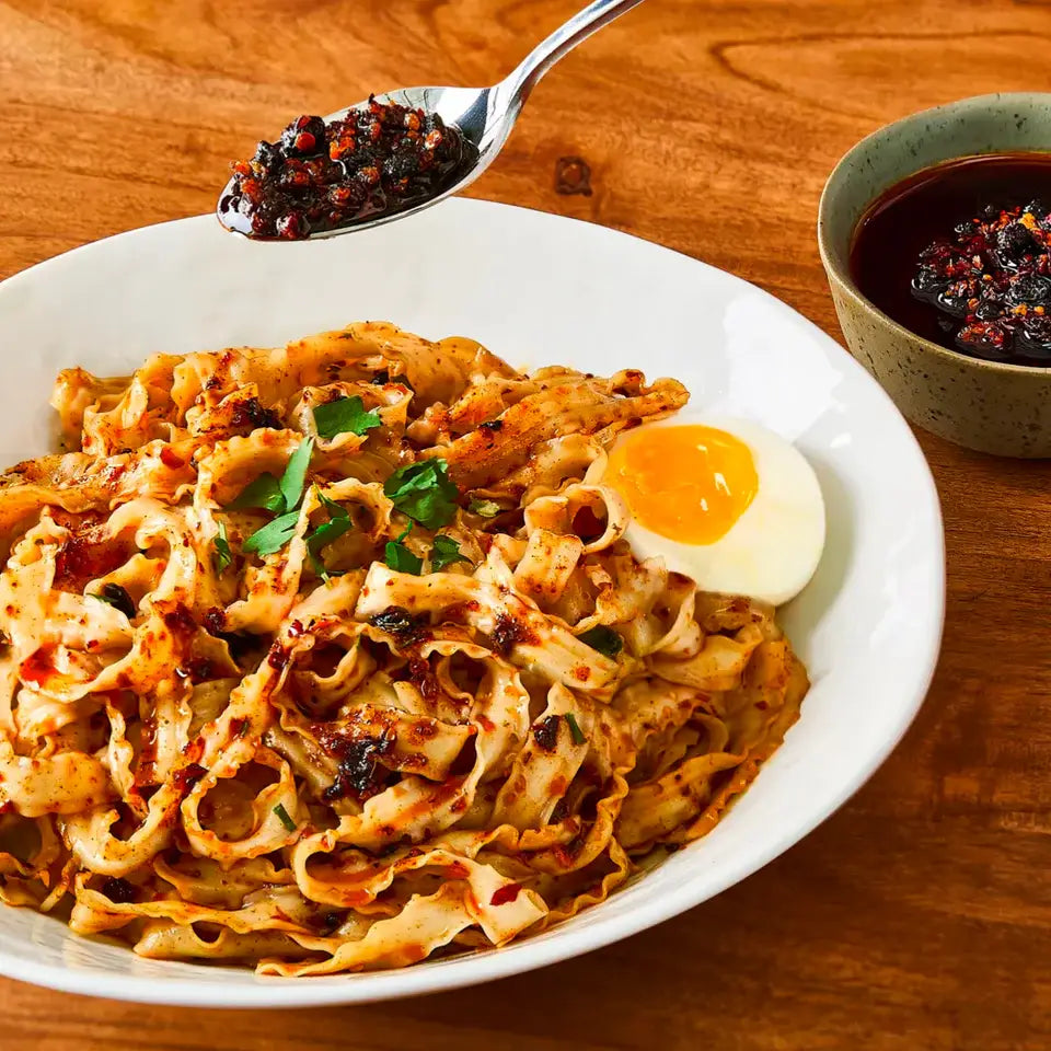 Pasta dish with a soft-boiled egg and a side of red pepper flakes on a wooden table.
