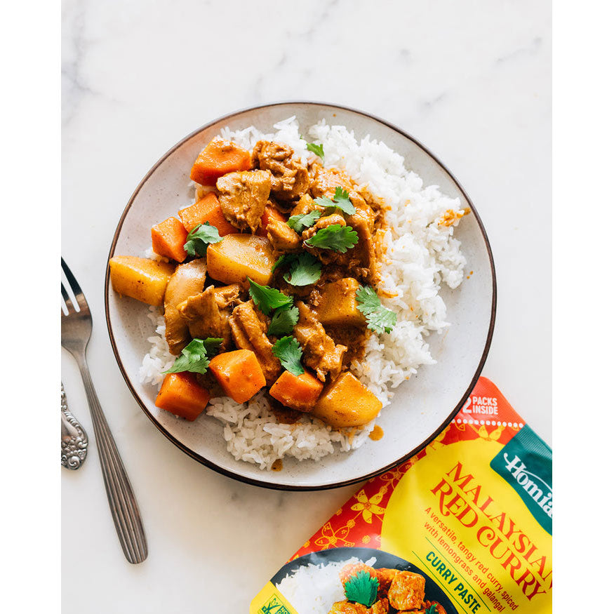 Plate of curry with rice and sweet potatoes next to a packet of Homdox Malaysian Red Curry Paste on a marble surface.