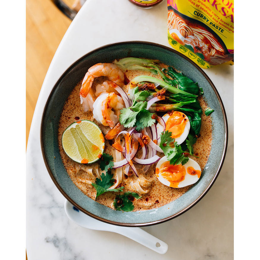  bowl of noodles with shrimp, lime, and egg on a marble surface with curry paste container.