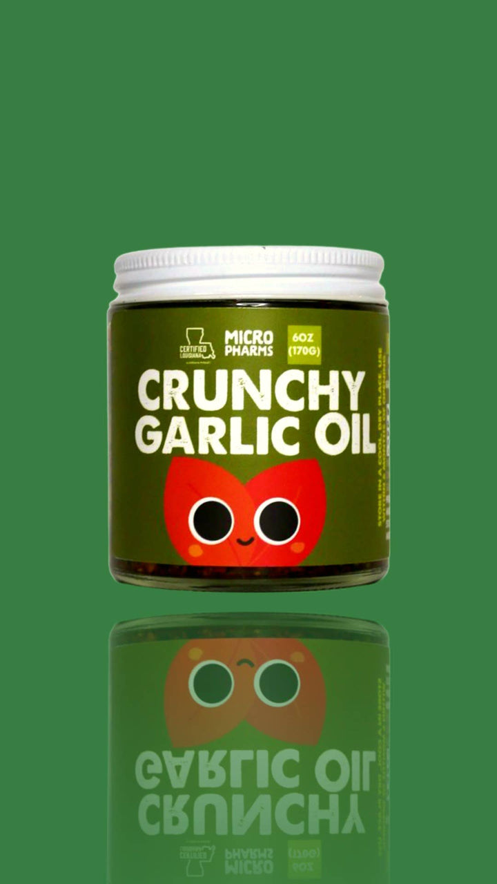 A jar of Micro Plumps Crunchy Garlic Oil centered on a solid green background with a reflection below. The jar has a white lid, a dark olive green label with bold white text, and a red cartoon garlic bulb graphic.