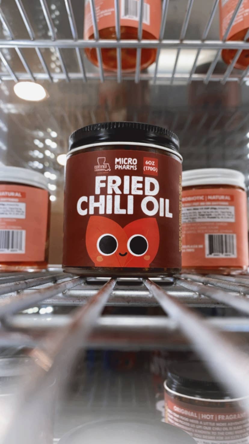 A jar of Micro Pharyngis Fried Chili Oil displayed on a metal grocery shelf, with other jars blurred in the background.
