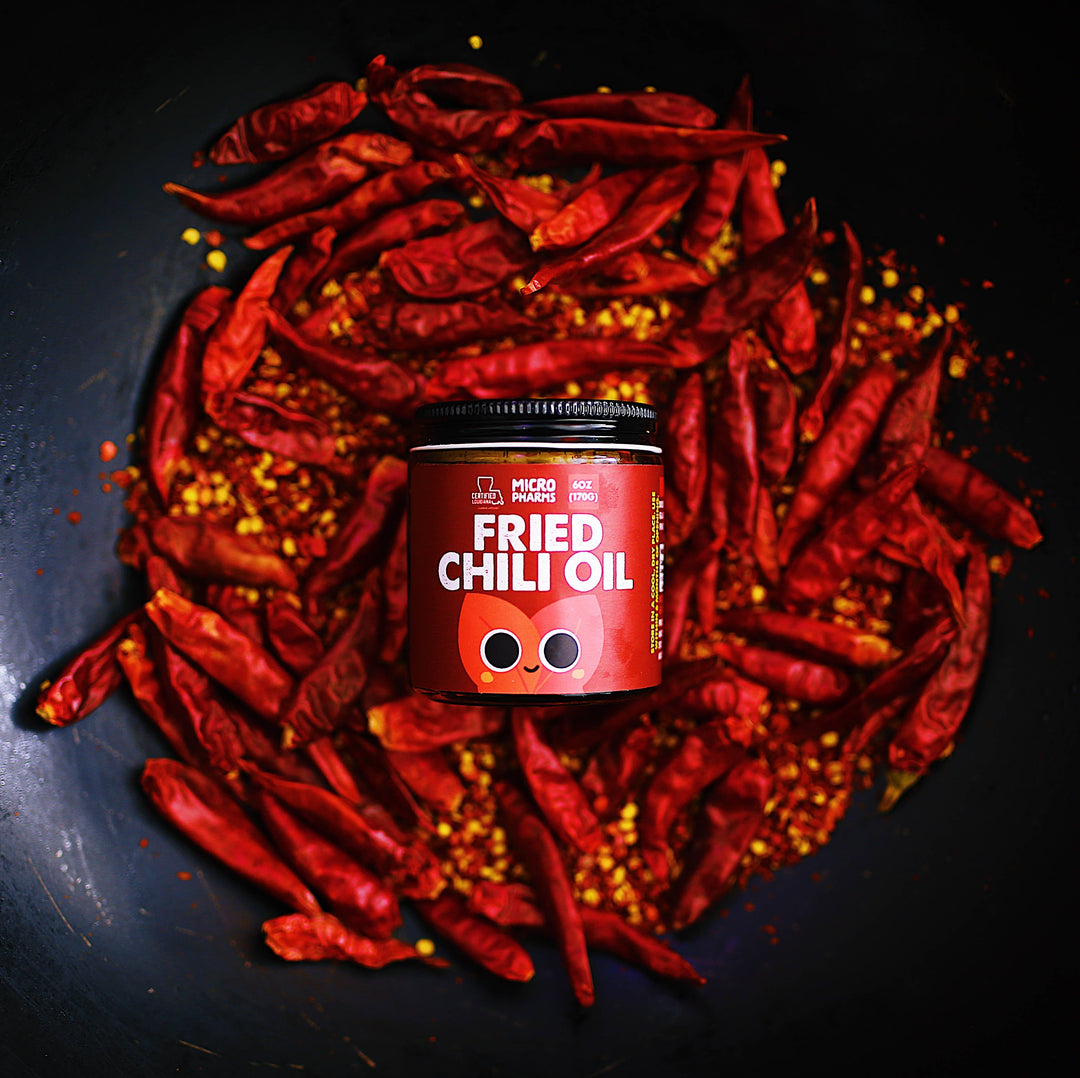 A jar of Micro Ingredients Fried Chili Oil placed in the center of a dark pan filled with dried red chili peppers and chili flakes.