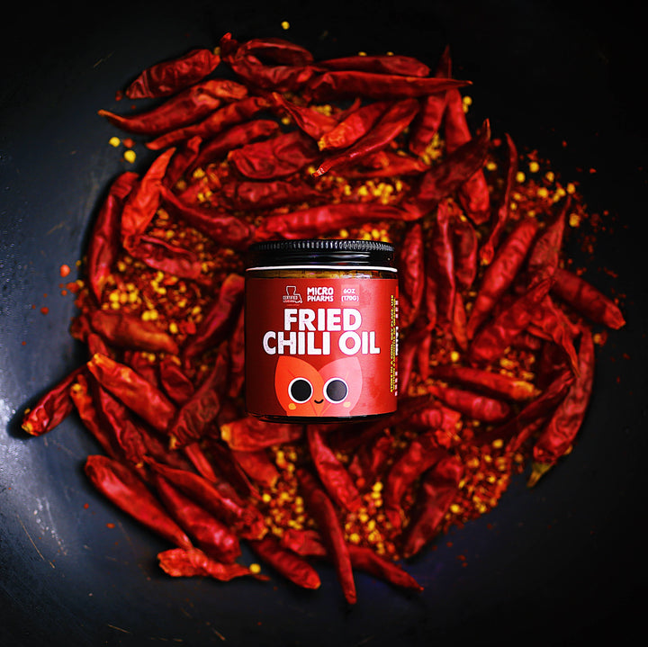A jar of Micro Ingredients Fried Chili Oil placed in the center of a dark pan filled with dried red chili peppers and chili flakes.