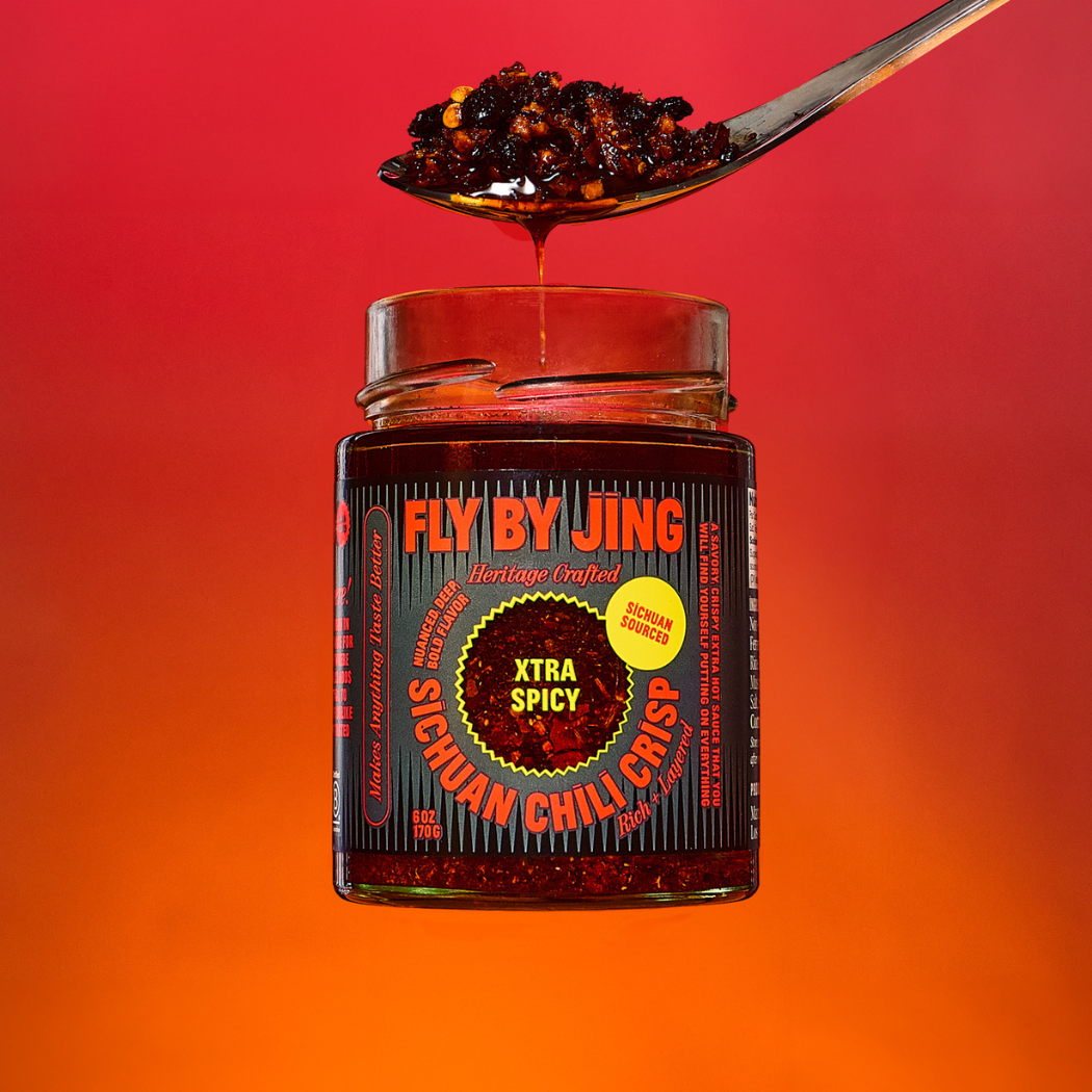 Jar of 'Fly By Jing' Xtra Spicy Sichuan Chili Crisp with a spoonful of chili on a red and orange gradient background.